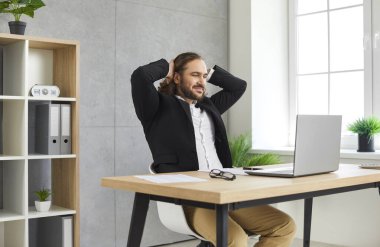 Genç, kendine güveni tam, uzun saçlı iş adamı bilgisayar ekranına bakarak ofiste dizüstü bilgisayarla çalışıyor. İşyerindeki masada oturan memnun adam..