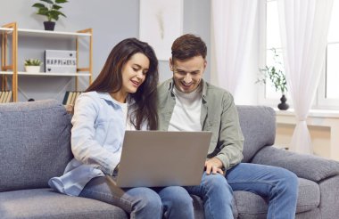 Genç bir aile, modern oturma odasındaki kanepede oturmuş dizüstü bilgisayar ekranına bakan sevimli bir çift. Mutlu çift, evli eş web sörfü, online alışveriş yapmak veya bilet ayırtmak, video izlemek