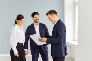 Genç evli çift yeni bir ev, daire ya da daire alıyor. Mutlu aile, karı koca, beyaz oturma odasında bir araya gelip emlakçıyla ya da emlakçıyla kontrat imzalamaya hazır.