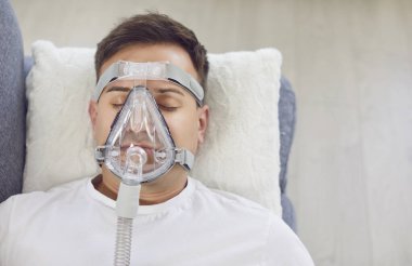 Zatürree ya da bronşiti olan genç bir adamın yüzünde tıbbi oksijen solunum maskesi vardı. Evdeki kanepede, yumuşak beyaz bir yastığın üzerinde yatıyordu. Sağlık hizmeti, oksijen terapisi konsepti