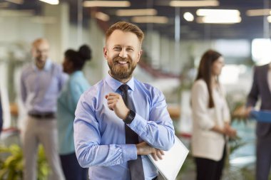 Resmi giysiler içinde gülümseyen, kendine güveni tam genç bir iş adamının portresi. Ofiste çalışan şirket çalışanlarından oluşan bir ekiple kamera karşısında neşeli görünüyor. İş portresi kavramı