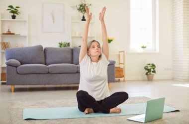 İnternetteki eğitim sırasında yerde dizüstü bilgisayarla yoga yapan rahat yaşlı kadın portresi. Emekli kadın evde meditasyon yapıyor. Emeklilik konseptinde spor.