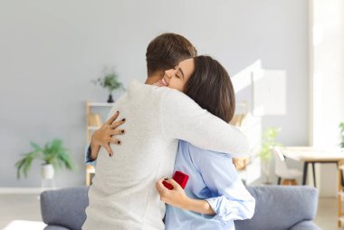 Sevgili çift, son nişan tekliflerini kutlarken sıkıca sarılıyor. Kadının elinde nişan yüzüğü için küçük kırmızı bir kutu var. Yüzündeki ifade mutluluk ve sevgi saçıyor..