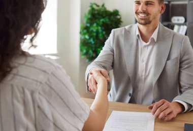 Business woman sitting in office on workplace shaking hands with smiling cheerful man colleague reaching agreement making a good deal or signing a contract. Business people at work.
