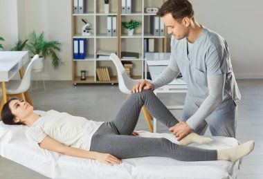 Erkek fizyoterapist, rehabilitasyon sırasında kadın hastanın bacağını esnetmesine yardım ediyor. Klinikte kanepede uzanan genç bir kadın yaralanma tedavisi ve hareket desteği görüyor..