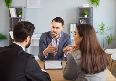 Erkek mali danışman, ofisteki masada oturan kendine güvenen genç bir çifte sigorta danışmanlığı yapıyor. Kadın ve erkek emlakçıyla ya da komisyoncuyla konuşuyor..