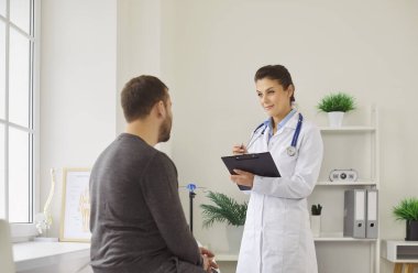 Beyaz üniformalı kadın doktor erkek hastayı açıklıyor, sağlık tedavi sonuçları hakkında iyi haberler veriyor, hastalıkları tedavi ediyor, sağlık kontrolünde motive ediyor ve cesaretlendiriyor, yardım ediyor, yardım ediyor.