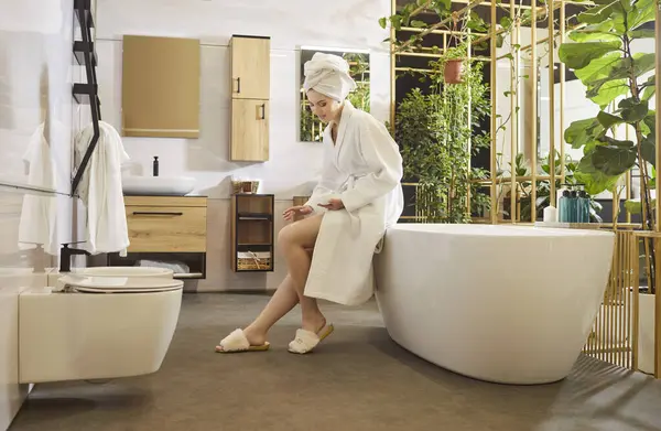 Kafasına beyaz bornoz ve havlu geçirmiş rahat bir kadın, banyo yaptıktan sonra bacaklarına krem sürüyor, küvette modern iç mekanlara karşı poz veriyor, günlük güzellik ve vücut bakımı prosedürleri uyguluyor.