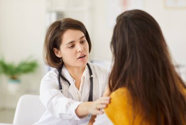 Beyaz önlüklü, şefkatli bir kadın doktor parlak bir muayenehanede oturuyor ve nazikçe hastayı rahatlatıyor. Bakım hekimi, sağlık danışmanlığı sırasında genç bayana destek ve anlayış sunuyor.