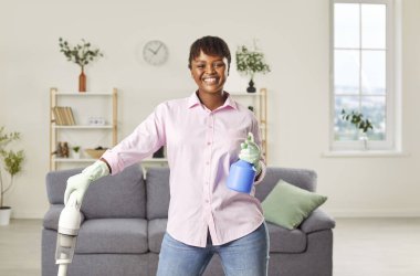 Gülümseyen mutlu Afro-Amerikan kadın elinde elektrikli süpürgeyle evdeki oturma odasında neşeli görünüyor. Temizlik, temizlik, ev işleri ve ev işleri.