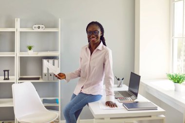 Mutlu ofis çalışanı, sekreter, muhasebeci ya da işletme müdürünün portresi. Sırıtkan Afrikalı genç bir Amerikalı kadın elinde evrak, dizüstü bilgisayar, hesap makinesi ve kağıtlarla masa başında duruyor.