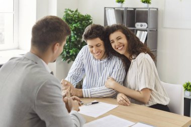 Ofisteki emlakçıyla görüşürken birbirine yakın oturan ve kucaklaşan mutlu genç çift. Gayrimenkul alımını ya da emlak sözleşmesini imzaladıktan sonra kirayı kutlayan neşeli erkek ve kadın..