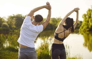 Aktif çiftlerin spor eğitimi, kadın ve erkek sabah egzersizleri sırasında vücut esneme hareketleri. Kolları havada atletlerin göl kenarında yan yana eğilmelerini izliyoruz. Sağlıklı egzersiz, fitness konsepti