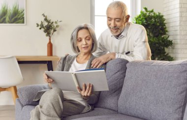 Yaşlı çift mutlu mesut aile fotoğrafı albümüne bakıyor, evdeki kanepede oturuyorlar. Neşeli aktivite, aile anılarının boş zamanlara mutluluk getirmesinin önemini vurgular..