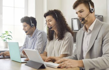 Genç iş adamları çevrimiçi destek servisinin çağrı merkezinde sıraya girip çalışıyor. Kulaklık takmış operatör ajanları yardım masasında dizüstü bilgisayarla çalışan müşterilerle iletişim kuruyor..