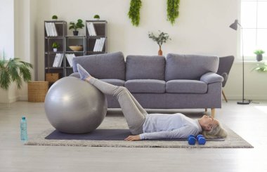 Yaşlı bir kadın yoga minderinde yatarak spor egzersizleri yapıyor. Olgun bir kadın evdeki oturma odasında egzersiz yapıyor. Sağlıklı yaşam tarzı, fitness ve spor kavramı.
