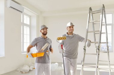 Üniformalı iki gülümseyen erkek ressam ellerinde boya ruloları ve tepsiler merdivenin yanındaki parlak odada duruyorlardı. Mutlu işçiler yenileme ve duvar boyama için hazırlanıyor, iç tasarımı yenilemeye hazır..