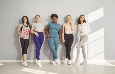 Yoga yaptıktan sonra duvarda poz veren mutlu, çok ırklı bir grup genç kız. Spor salonu, spor salonu, spor salonu, zayıf arkadaşlar.