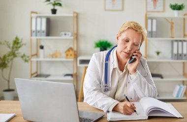 Hastane ofisinde dizüstü bilgisayarla çalışırken telefonla konuşan konsantre yaşlı doktor ya da hemşire. Sağlık, internet bağlantısı ve mesleki adanmışlık..