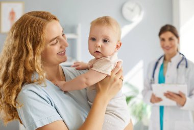 Mutlu gülümseyen genç bir annenin portresi. Bebek kadar tatlı bir bebek. Klinikte duran çocuk doktoru arkadaşını ziyaret ediyor. Sağlık hizmeti kavramı.
