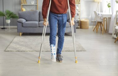 Evdeki oturma odasında koltuk değneği kullanarak kırık bacağı olan engelli bir adamın kesilmiş fotoğrafı. Bacağı alçılı özürlü adam. Rehabilitasyon ve iyileşme kavramı.