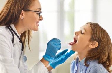 Kadın gülümseyen çocuk doktorunun sağlık kliniğinde küçük erkek hastaları muayene edişinin yan görüntüsü. Doktor kadın otolarolog muayenehanede hasta bir çocuğun ağız boşluğunu muayene ediyor..