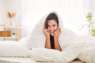 Yüzünde gülümseme olan genç bir kadın, yumuşak yastığa uzanmış, rahat bir yatakta, başını sıcak, beyaz bir battaniyeyle. Rahatlamış mutlu esmer kız evde huzurlu bir sabahın rahatlığının ve huzurunun tadını çıkarıyor..