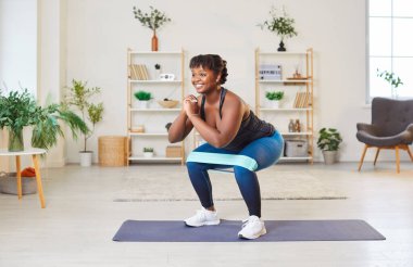 Spor formundaki orta yaşlı mutlu Afro-Amerikalı kadın oturma odasında elastik bantla egzersiz yapıyor spor kıyafetleri giyip sağlıklı bir yaşam tarzı için evde egzersiz yapıyor.