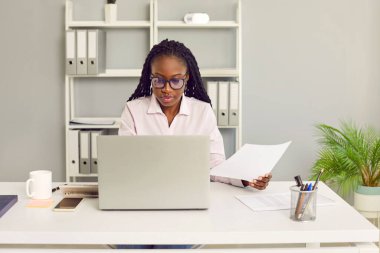 Yoğunlaştırılmış Afro-Amerikan iş kadını bir muhasebecinin portresi. İş yerindeki bir gömlekte oturuyor ve ofiste bir dizüstü bilgisayar üzerinde çalışıyor. Monitör ekranına bakıyor..