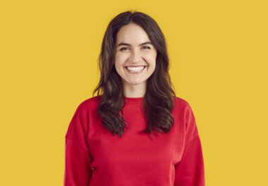 İnsanlar ve samimi duygular. Yüzünde dişlek bir gülümseme olan pozitif esmer kadın gündelik kırmızı kazak giyer, parlak sarı arka plana karşı poz verir, mutluluğu ifade eder, iyi bir ruh hali içindedir.