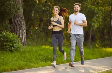 Genç, gülümseyen mutlu aktif çift spor eğitimleri sırasında parkta birlikte koşuyorlar. Erkek ve kadın dışarıda koşuyor. Doğada egzersiz ve sağlıklı yaşam tarzı konsepti.