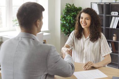 Pozitif kadın, ofiste masada oturmuş işbirliği için uzlaşmaya varırken bir erkekle tokalaşıyor. Kıvırcık saçlı kadın model iş görüşmesine geldi, çalışmaya hazır.