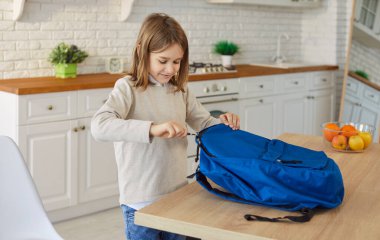 Küçük öğrenci eşyalarını mavi sırt çantasına koyuyor. Okuldan önce ya da öğrenim etkinliği düzenliyor. Modern mutfak ortamında duruyor.