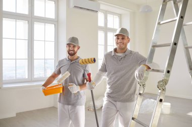 Ev dekorasyonu için boya malzemesi üreten iki mutlu profesyonel duvar ressamı. Üniformalı gülümseyen erkek işçiler merdiven başında, ellerinde boya tepsisi ve rulo tutuyorlardı.
