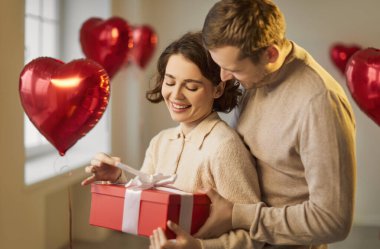 Sevgililer günü hediye kutusu hediye paketi vermek, aşk hissi, romantik sürpriz, sevgi dolu bir çift şefkatle kucaklaşmak, öpüşmek, erkek, kadın, kalp şeklinde kırmızı balon folyo dekoru, düğün odası dekorasyonu, evlenme teklifi etmek.