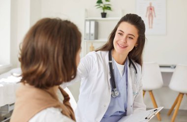 Beyaz önlüklü gülümseyen doktor, parlak tıp ofisinde genç bir bayanın yanında oturuyor. Dost canlısı bayan kardiyolog hasta omzunda dinleniyor. Muayeneleri sırasında destek ve bakım öneriyor..