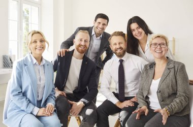 Başarılı iş ekibinin ve şirket çalışanlarının grup portresi ofis alanında yan yana oturup duruyor. Zeki takım elbiseli mutlu meslektaşlar gülümsüyor, grup fotoğrafı için poz veriyor, kameraya bakıyor..