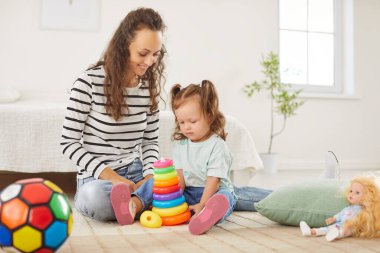 Anne ve çocuk birlikte vakit geçiriyorlar. Anne ve küçük kızı aydınlık odada halıya oturmuş birlikte oynuyorlar. Küçük kız çok renkli oyuncak piramidine dikkatle bakıyor..