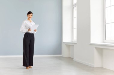 Kendine güvenen genç bir emlakçının portresi yeni boş dairelerde elinde belgelerle kameraya neşeli bir şekilde bakıyor. Emlak piyasası ve taşınma günü konsepti.