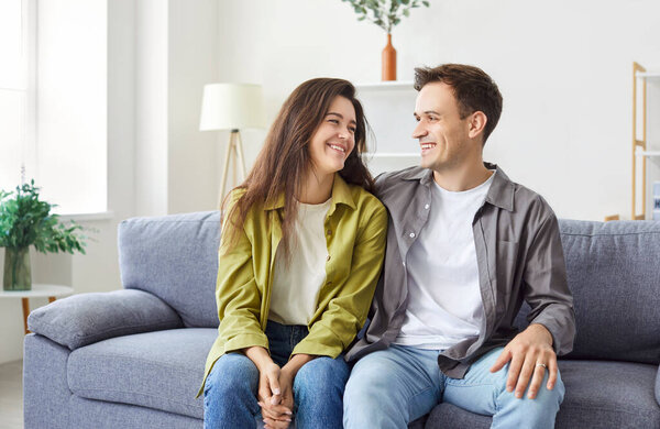 Портрет молодой счастливой супружеской пары, сидящей вместе на диване, обнимающейся и смотрящей друг на друга. Веселый улыбающийся молодой кавказский мужчина и женщина на диване в гостиной, излучающей любовь и счастье.