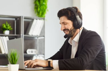 Çağrı merkezi operatörü kulaklıkla masa başında oturuyor. Laptop üzerinde çalışıyor. Müşteri hizmetleri için ofisteki müşterilerle iletişim kuruyor. Teknik destek ve büro kavramına yardım.