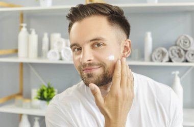 Erkekler için cilt bakımı. Cilt bakımını seven bir adam banyoda gülümseyerek nemlendirici sürüyor..