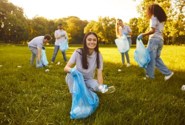 Eldivenleri ve çöp torbasıyla mutlu genç bir kadının portresi. Arka planda bir gönüllü ile yaz parkında plastik çöp topluyor. Çevre kirliliği ve ekoloji kavramı.