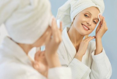 Kafasına havlu sarılmış yumuşak bornoz giymiş neşeli bir kadın nazikçe losyon veya toner sürüyor. Mutlu gülümseyen kadın aynadaki yansımasına bakarken günlük yüz bakımından zevk alıyor..