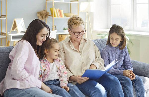 Happy family day at home, granny older woman, mother, two kids sitting on sofa, reading book together, looking and smiling, enjoying shared storytelling time on weekend, connected, interacting 
