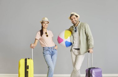 Mutlu çift yaz tatiline çıkmaya hazırlanırken beklenti ve sevinçle doludur. Gri arka planda duran gündelik elbiseli çift plaj topu ve bavullarını tutuyorlar..