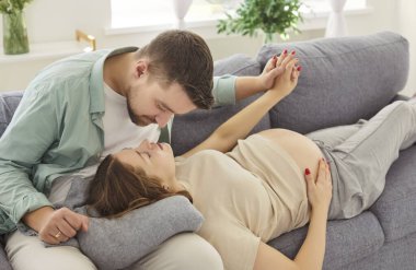 Oturma odasındaki kanepede oturan genç adam ve hamile karısı birbirine şefkatle bakıyor. Bebek bekleyen mutlu çift ebeveyn olmaya hazırlanıyor. Hamilelik ve ebeveynlik kavramı.