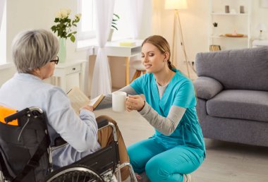 Genç, dost canlısı bayan sosyal hizmetler görevlisi tekerlekli sandalyede kitap okuyan yaşlı ve engelli bir kadına bir bardak içki veriyor. Huzurevinde yaşlı bir hastayla neşeli bir hemşire ya da bakıcı.