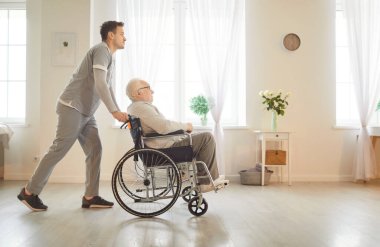 Üniformalı genç erkek hemşire, tıbbi klinikte tekerlekli sandalyedeki engelli yaşlı hastayı itiyor. Huzurevinde engelli, yaşlı bir adam. Bakıcı desteği kavramı.