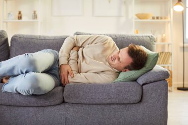 Hasta genç bir adam, acı dolu bir ifadeyle karnını tutarak kanepede yatıyor. Karın ağrısı, güçlü kramplar, rahatsızlık ya da hastalıktan muzdarip sağlıksız erkek, kıvrılıp yardım arıyor..